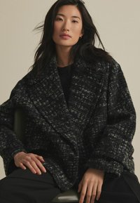 Üks suur must ja hall tweed mantel tekstuuriga mustriga, sakiliste krae ning nupuvõrudega. Loodud mugavaks sobivuseks, millel on üles keeratud varrukad.