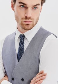 Szaro-biała kamizelka w kratę noszona na białej koszuli, w zestawie z granatowym wzorzystym krawatem. Kamizelka ma dekolt w serek oraz guziki.