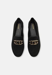 Mocassins en daim noir avec un accent de chaîne dorée portant un logo en forme de lettre 'G'. Texture lisse et design discret.