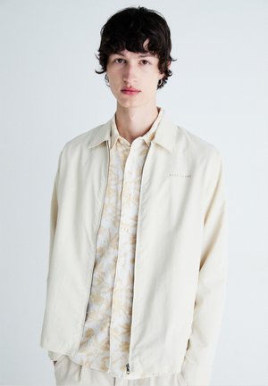 Chaqueta bomber de color beige claro sobre una camisa crema con un patrón floral. La chaqueta presenta un cierre de cremallera y un cuello clásico.