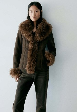 Jeune femme portant une veste marron avec un grand col et des poignets en fourrure, assortie à un pantalon foncé, debout devant un fond gris clair.