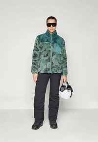 Person wearing green patterned fleece jacket, black pants, black boots, large dark sunglasses, holding white ski helmet by side.