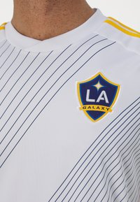Maglia sportiva bianca con strisce verticali blu, scollatura a girocollo, con logo ricamato della LA Galaxy in blu e giallo sul petto.