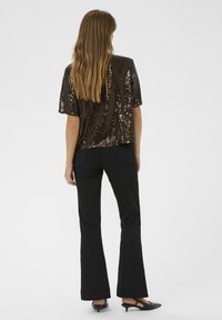 Vrouw met lang bruin haar draagt een kort-slauwen donkerbruine pailletten top, zwarte flared broek en zwarte kitten heel schoenen, met haar rug naar de kijker gekeerd.