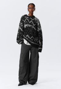 Pull en tricot noir et gris avec un motif géométrique, associé à un pantalon cargo noir oversize avec des poches latérales et des poignets ajustés.