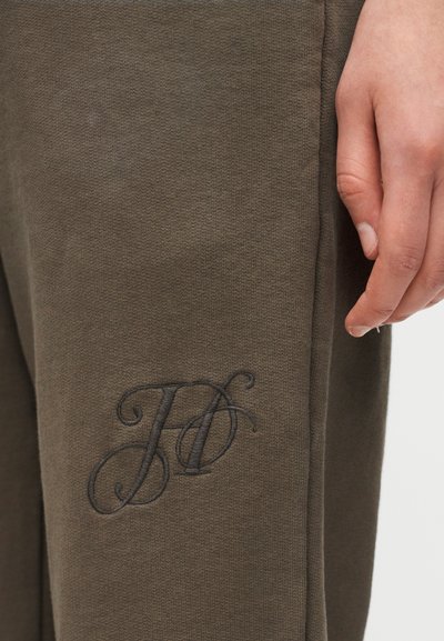 Χέρι που ακουμπάει δίπλα σε ύφασμα ελιάς με μαύρα κεντημένα διαπλεκόμενα γράμματα "J" και "D" κοντά στη ραφή.