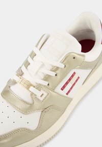 Baskets blanches et dorées avec une tige en cuir lisse et en mesh, dotées d'accents dorés, de perforations, de lacets blancs et d'un patch logo rouge.