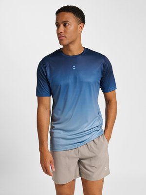 Man staat met linkerhand in zak, draagt een blauw verloop kortärmelig sportshirt en beige korte broek tegen een witte achtergrond.