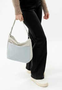 Bolso de hombro de cuero gris claro y crema con un diseño relajado, que presenta un bolsillo frontal en azul pálido y herrajes plateados.