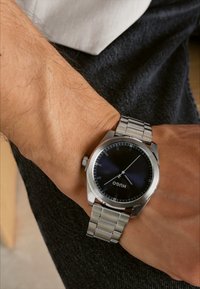 Reloj de acero inoxidable plateado con un dial azul oscuro, pulsera de eslabones y marcadores de horas sencillos. Presenta la marca "HUGO" exhibida de manera prominente.