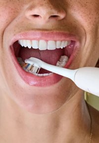 Brosse à dents électrique avec un manche blanc, des poils bleus et jaunes, dans une bouche affichant des dents blanches et propres ainsi que des gencives saines.