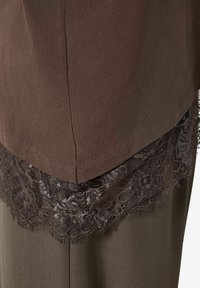 Brūns kokvilnas tops ar tumši brūnu mežģīņu apakšmalu ar ziedu rakstiem. Audums ir gluds, kontrastējot ar mežģīņu tekstūru.