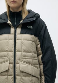 Μπεζ και μαύρο αδιάβροχο μπουφάν με υφή ματ, διαθέτει κλείσιμο με φερμουάρ, κουκούλα και δύο μπροστινές τσέπες. Λογότυπο στην αριστερή πλευρά του στήθους.