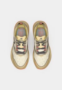 Chaussures outdoor beige clair avec dessus texturé, accents gris, lacets rouges et semelles rembourrées. Caractéristiques avec le logo "New Balance Hierro".