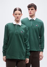 Δύο μοντέλα φορούν ταιριαστά πράσινα πόλο μπλουζάκια με μακρύ μανίκι και λευκό γιακά, μαζί με καφέ παντελόνια, κοιτώντας προς τα εμπρός μπροστά σε απλό φόντο.