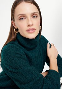 Il pullover a collo alto in maglia teal presenta una texture a coste e maniche ampie, abbinato a orecchini a cerchio dorati e un anello d'argento. 