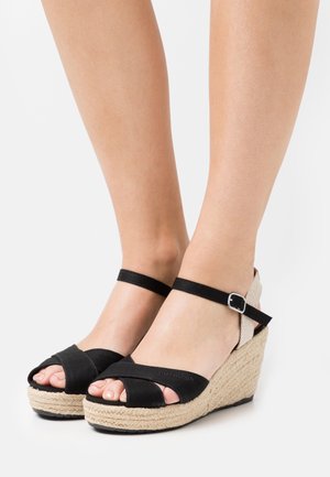 Sandales à talons compensés en tissu noir avec des brides croisées, semelle en jute, bride de cheville réglable et talon texturé de couleur claire.