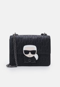 Musta nahkainen käsilaukku, jossa on painettu logokuvio, hopeinen ketjuhihna ja etutasku, jossa on aurinkolasit päässään oleva sarjakuvahahmo.