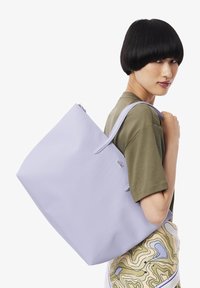 Bolso tote de color lavanda claro con cierre de cremallera, hecho de material texturizado. Cuenta con asas dobles y un pequeño logo decorativo.