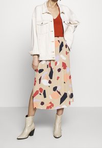 Farkkutakki, punainen toppi ja beige hame, jossa on abstrakteja monivärisiä kuvioita. Nilkkapituinen hame, jossa on sivuhalkio; kiiltävät beige cowboy-kengät.