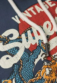 Sudadera azul marino con un estampado agrietado que presenta diseños de un dragón azul y un león naranja, cordones blancos y un extremo metálico con la marca.