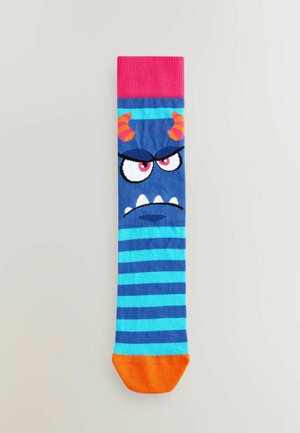 Modrá a tyrkysová pruhovaná ponožka s dizajnom príšery s rohmi, nahnevanými očami a oranžovým špičkou. Vrchná časť je rebrovaná a ružová.