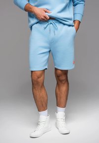 Shorts de survêtement bleu clair avec taille élastique et cordon de serrage, dotés d'une poche latérale et d'un petit détail logo rouge.