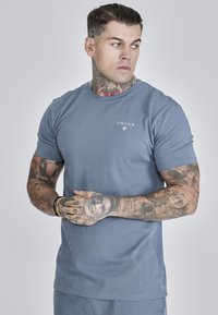 Tricou din bumbac albastru, cu guler rotund, mâneci scurte și un mic logo alb pe partea stângă a pieptului. Textură netedă, potrivire lejeră.