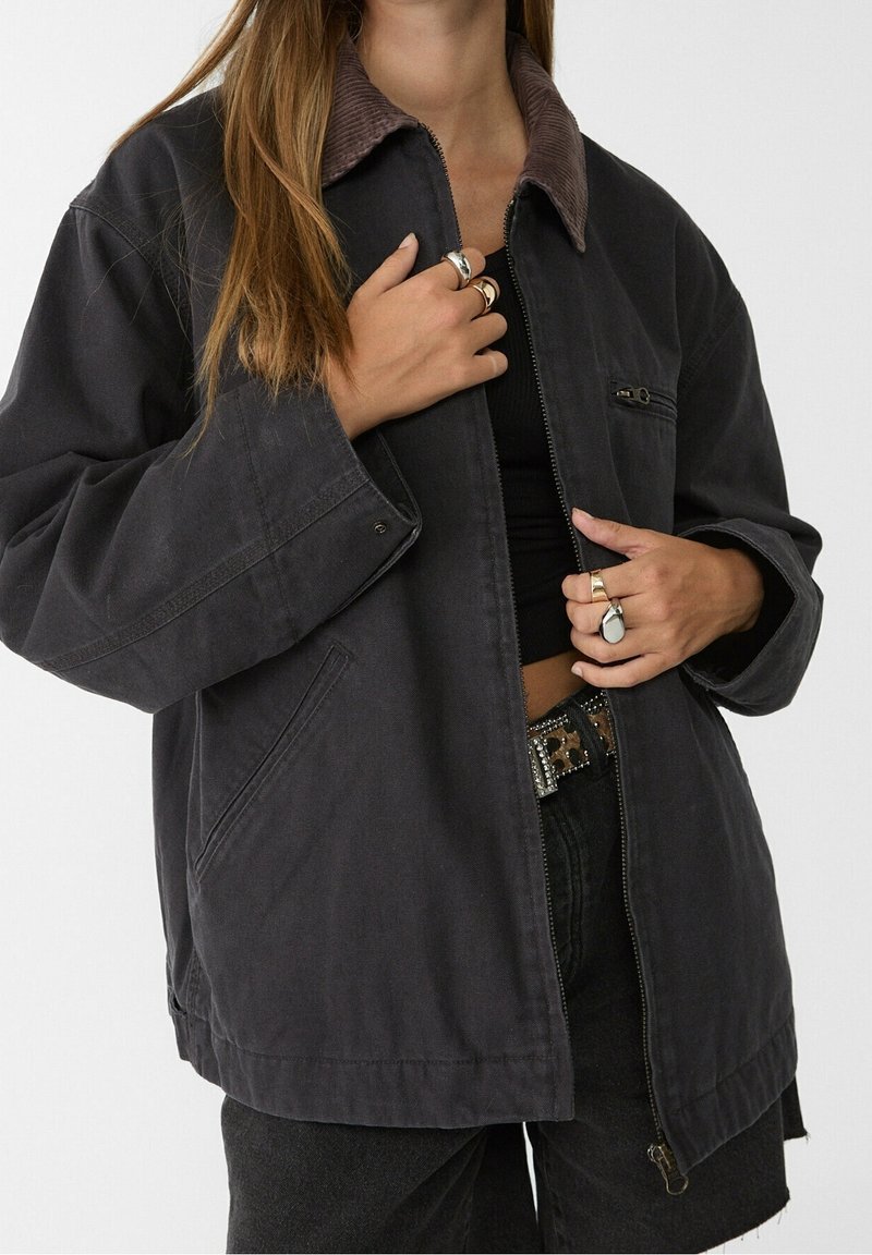 Oversized dark gray jacket with a corduroy collar, zip closure, side pockets, and a relaxed fit. Fabric appears sturdy and textured.
