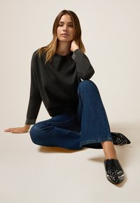 Maglione grigio con polsini strutturati, jeans blu a zampa d'elefante e scarpe nere con punta a stiletto decorate con borchie argentate. Posizione seduta su uno sfondo chiaro.