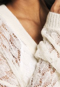 Bílé pletené cardigan s volným, texturovaným krajkovým vzorem a širokým V-výstřihem, se žebrovanými detaily na okrajích a měkkou, lehkou tkaninou.