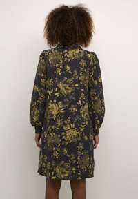 Mujer con cabello rizado vestida con un vestido negro con patrones florales amarillos, de pie frente a una pared clara y sencilla.