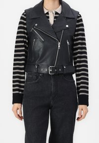 Person, der eine schwarze Lederweste ohne Ärmel mit Reißverschluss und Gürtel über einem schwarz-weißen gestreiften Langarmshirt und schwarzen Jeans trägt.