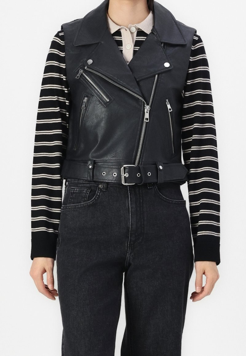 Person, der eine schwarze Lederweste ohne Ärmel mit Reißverschluss und Gürtel über einem schwarz-weißen gestreiften Langarmshirt und schwarzen Jeans trägt.