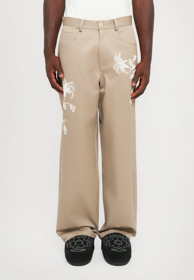 Beige weite Hosen aus glattem Stoff, mit weißer floralem Stickerei an beiden Beinen, ergänzt durch schwarze bestickte Schuhe.