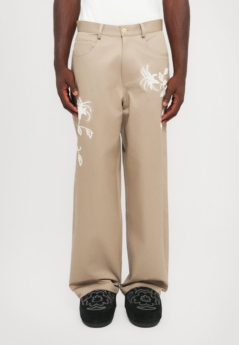 Beige vidde bukser lavet af glat stof, med hvid blomstermotiver broderet på begge ben, suppleret af sorte broderede sko.