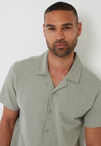 Camisa polo de manga corta, de color verde claro, con cuello, cinco botones y una tela texturizada. El modelo muestra la camisa frente a un fondo neutral.