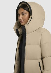 Giacca a vento beige con ampio cappuccio, dettagli neri e zip frontale. Presenta sezioni trapuntate per l'isolamento e regolazioni con cordoncino.