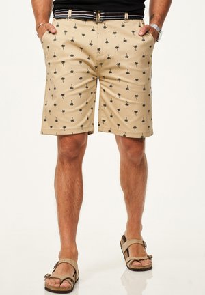 Mann trägt beige Shorts mit schwarzem Palmenmuster, gestreiften Gürtel, schwarzes Top und beige Sandalen, Hände in den Taschen, steht mit gespreizten Beinen.