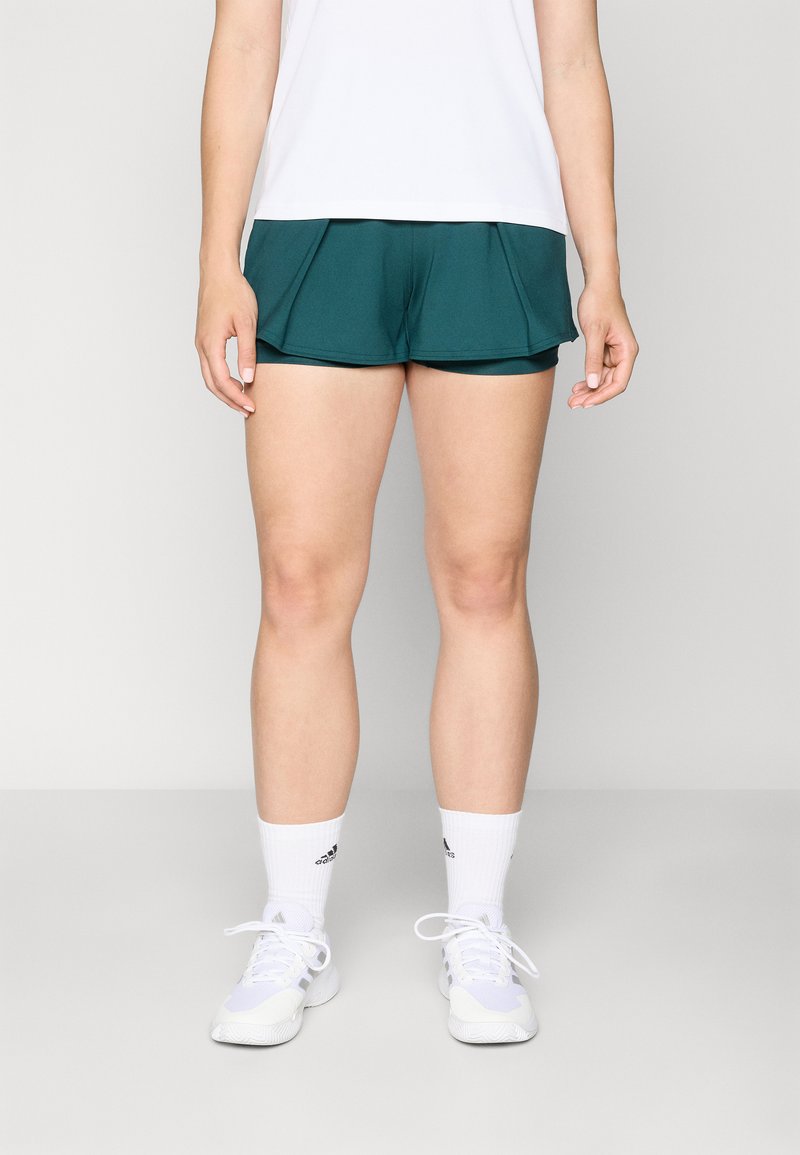 Shorts de sport teal en tissu respirant, avec un design en couches. Assortis à un haut blanc et des chaussures de sport blanches avec des accents noirs.