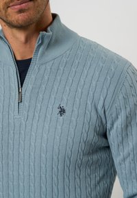 Šviesiai mėlynas megztinis su ketvirtį užtrauktuku, su motyvuotas kabelių raštu ir maža logotipu ant krūtinės. Minkšta tekstūra.