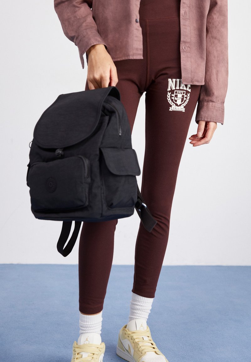 Black fabric backpack with rounded flap, front zip, and pocket. Worn with maroon leggings featuring a white logo, paired with yellow sneakers.