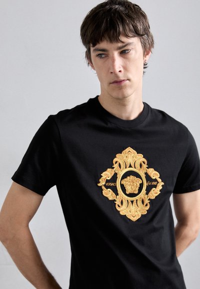 T-shirt en coton noir ornée d'un logo Versace brodé en or dans un design circulaire décoratif sur le devant. Manches courtes, col rond.