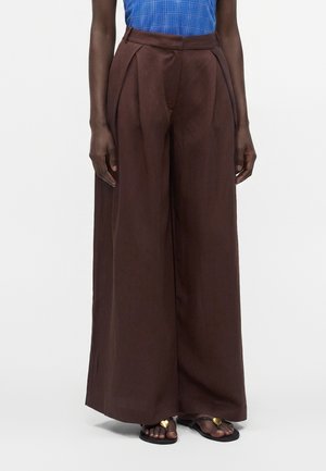 Personne portant un pantalon marron taille haute à jambes larges et des sandales noires avec une décoration dorée en forme de cœur, montrée de la taille aux pieds.