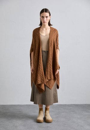 Brauner gestrickter Umhang mit Zopfmuster, getragen über einem beigen Oberteil und einem olivgrünen Rock. Das Model steht auf einem grauen Boden mit beigen Slippern.