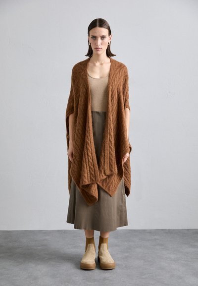 Avvolgente in maglia marrone con motivo a trecce, indossato sopra una maglietta beige e una gonna oliva. La modella è in piedi su un pavimento grigio con delle scarpe beige senza lacci.