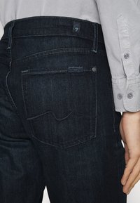 Jeans din denim de un albastru închis, cu o croială clasică, având un buzunar posterior cu detalii de cusătură și un logo subtil deasupra buzunarului. Materialul este neted.