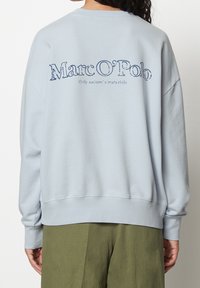 Persona che indossa una felpa azzurro chiaro con la scritta "Marc O'Polo" e "Only nature's materials" sul retro, abbinata a pantaloni verde oliva.
