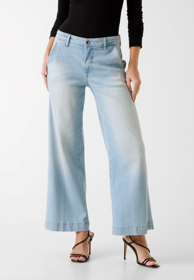 Γυναίκα που φοράει ανοιχτό μπλε τζιν με φαρδιά μπατζάκια, μαύρη μπλούζα με μακριά μανίκια και μαύρα ψηλοτάκουνα σανδάλια με λουράκια, στέκεται μπροστά σε λευκό φόντο.