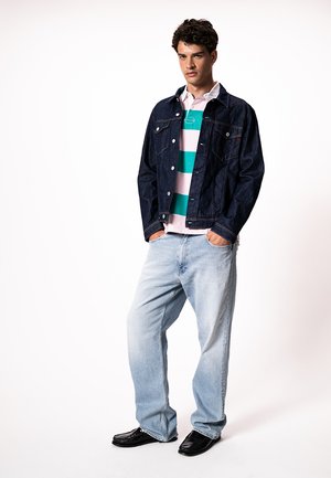 Ung mand står med hænderne i lommerne på lyseblå jeans, iført en mørk denimjakke, striped polo shirt og sorte sko på hvid baggrund.